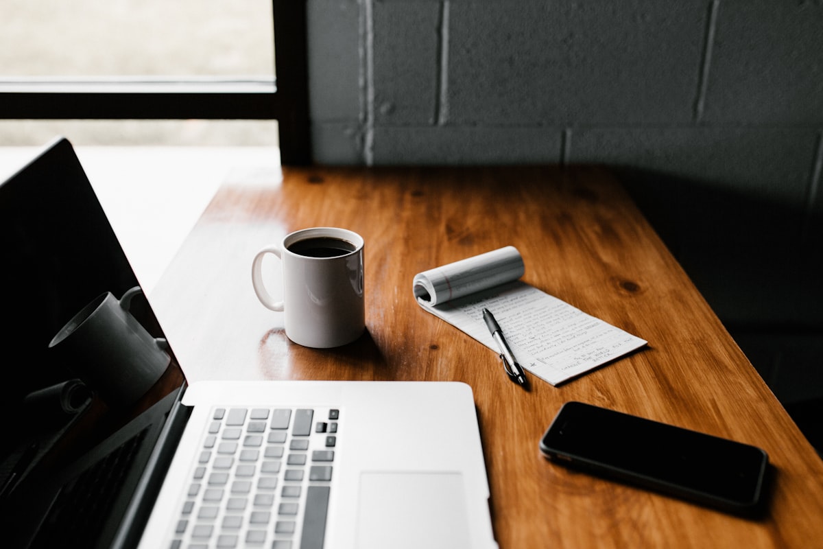 Laptop, coffee cup, notepad and phone on a wooden desk beside a window
