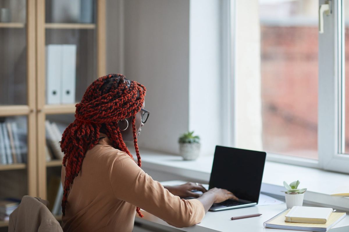 A practice owner with red braids and glasses working on a laptop at a desk by a sunny window